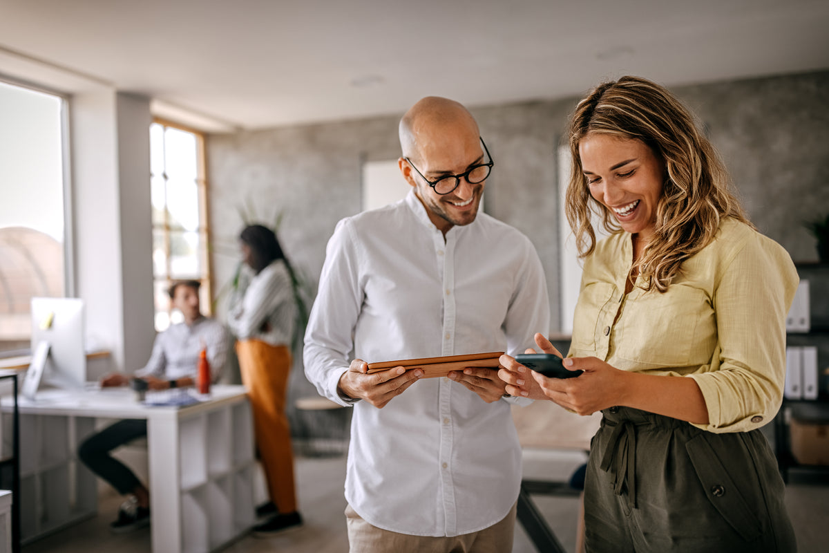Two colleagues smiling and looking at a phone together in a modern office setting.