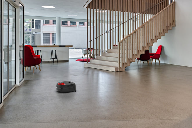 A robotic vacuum cleaner moves across the floor in a modern, open office with stairs and red chairs.