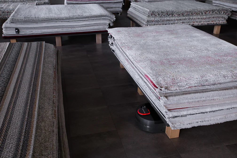 A robot vacuum cleans the floor under stacks of rugs in a showroom.