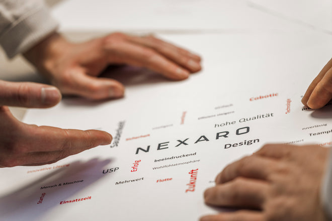 Close-up of hands pointing at a paper with the word NEXARO and other German words scattered around.