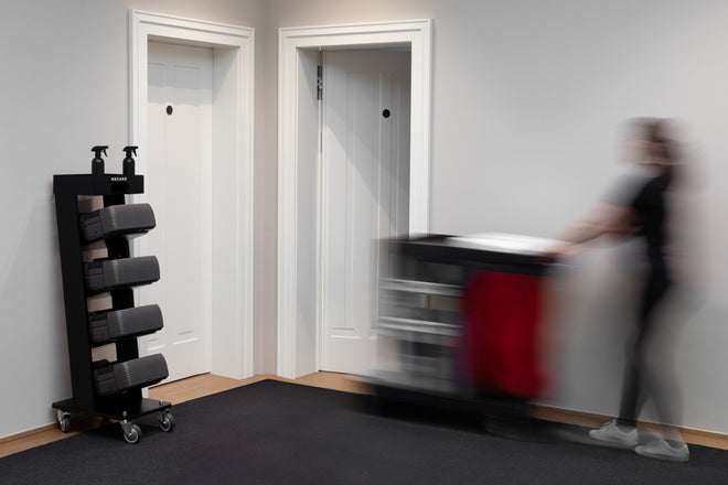 A blurred person pushes a cleaning cart past closed white doors in a hallway with a sanitizer stand.