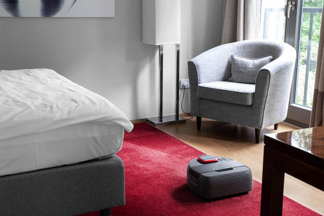 Ein Nexaro Saugroboter auf einem roten Teppich in einem Hotelzimmer mit Bett und Sessel.