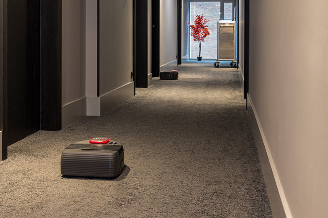 Two robotic suitcases move down a carpeted hotel hallway with a housekeeping cart in the distance.