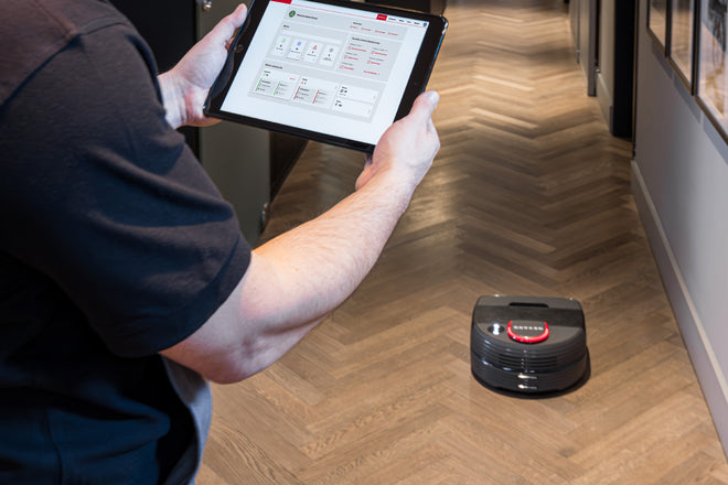 Person controlling a robot vacuum on a herringbone floor using a tablet with a smart home app.