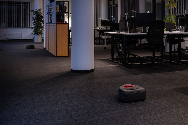 Two robot vacuums clean the floor in a modern, dimly lit office with desks, chairs, and plants.