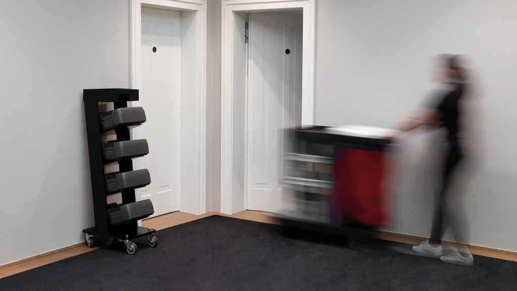 A person pushes a Nexaro trolley from Nexaro past two white doors and a black towel rack in a hallway, thereby increasing the efficiency of the cleaning team.