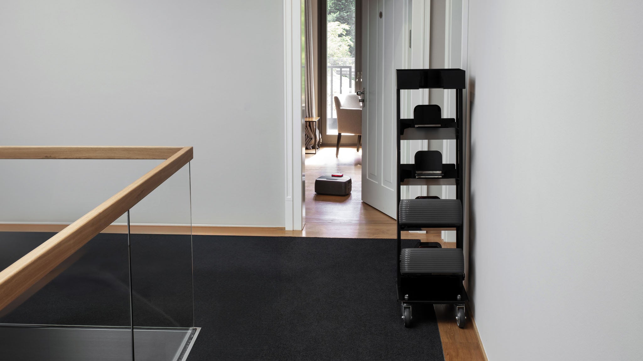 A black Nexaro trolley from Nexaro stands in front of a white wall in a modern hallway with carpeted and wooden floors, improving the efficiency of the transport and cleaning team.