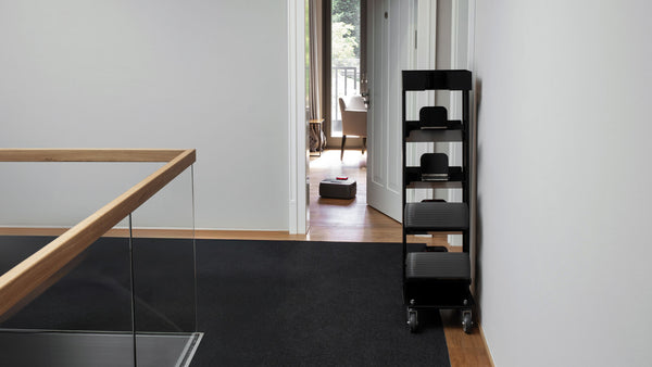 A black Nexaro trolley from Nexaro stands in front of a white wall in a modern hallway with carpeted and wooden floors, improving the efficiency of the transport and cleaning team.