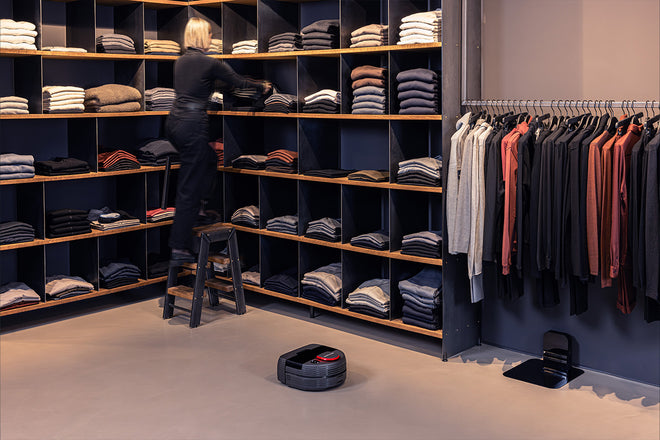 A sales assistant is sorting the clothes on a shelf. Next to her, a Nexaro robot vacuum cleaner is visible.