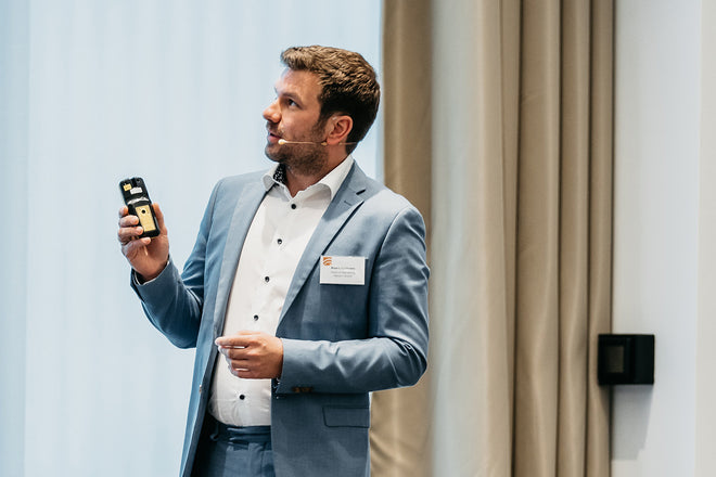 Man in a blue suit giving a presentation, holding a device and wearing a headset microphone.