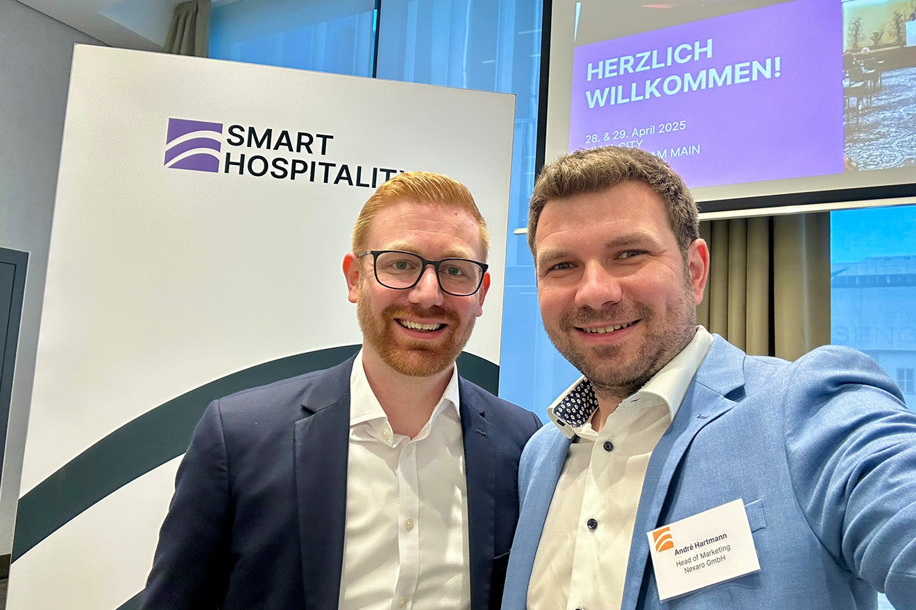 Two men in business attire smiling at a conference with a Smart Hospitality sign and welcome screen.