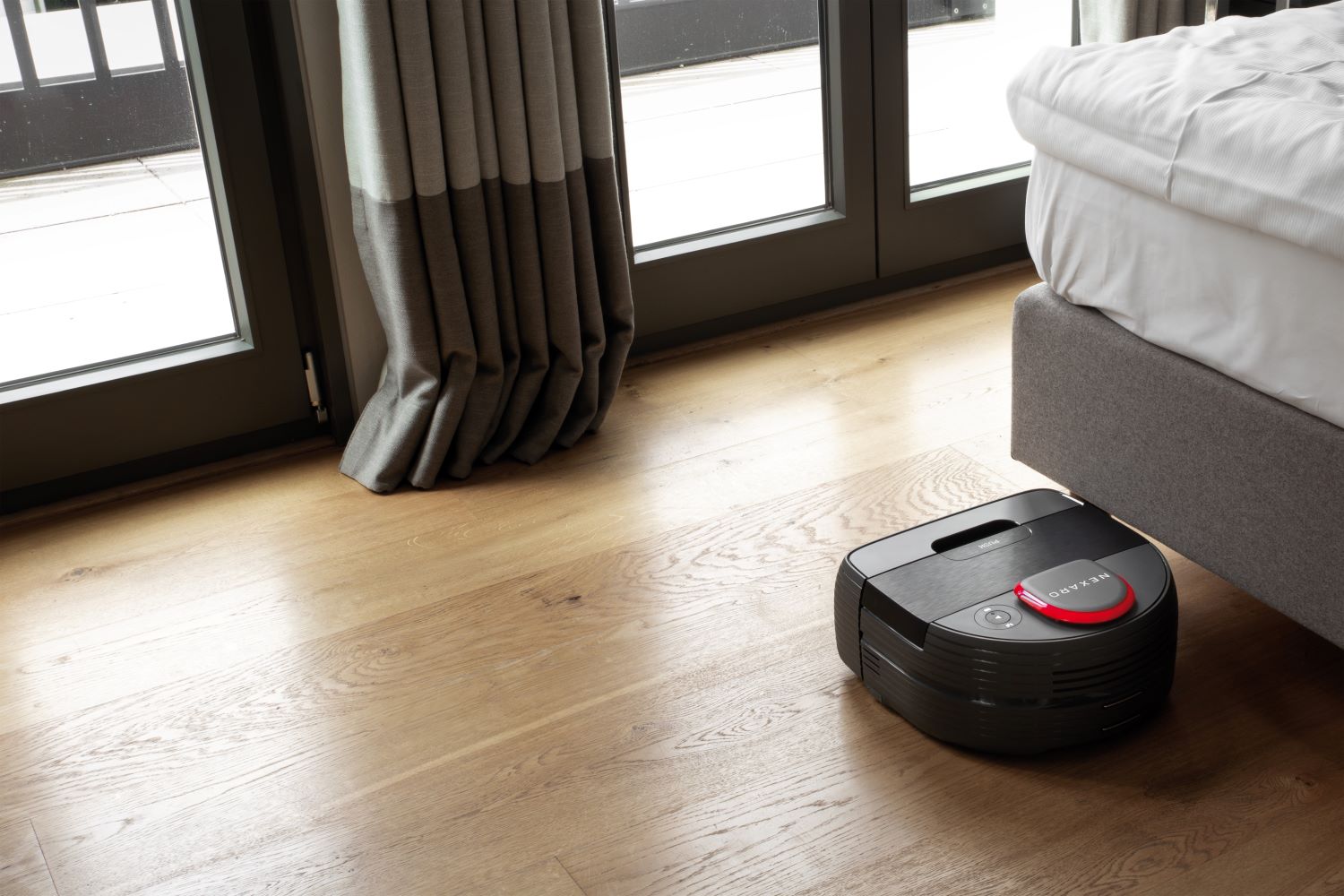 A robotic vacuum cleaner is cleaning a wooden floor next to a bed and large windows with curtains.