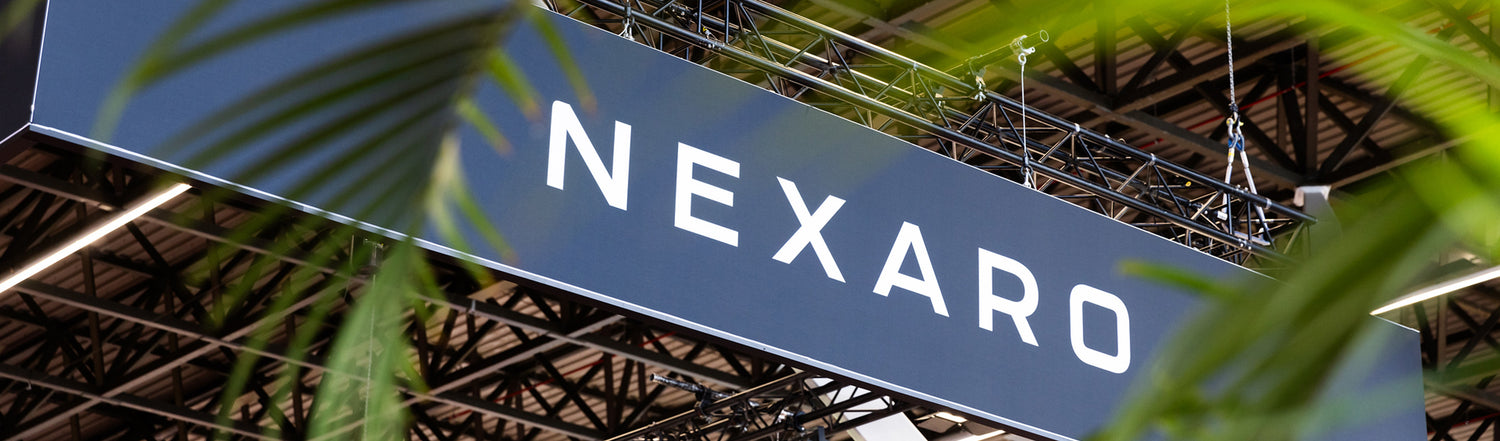 Large sign with the word NEXARO hangs from a ceiling, partially framed by green leaves in the foreground.
