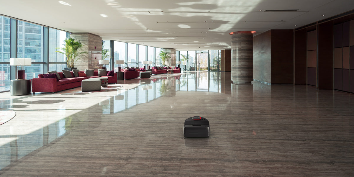 A robot vacuum cleans a spacious, modern lobby with large windows, red sofas, and potted plants.