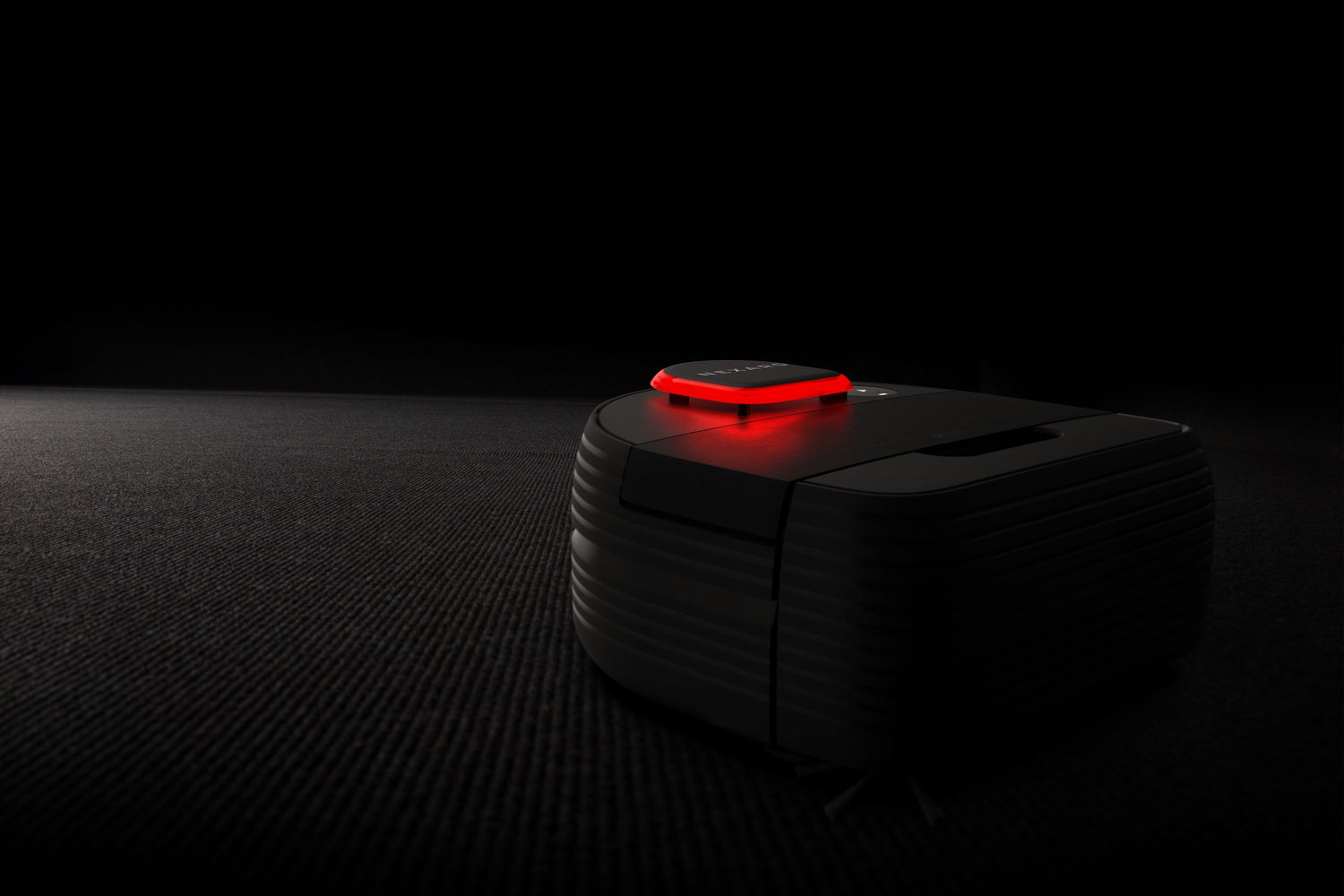 A black robot vacuum with a glowing red light sits on a dark textured floor in a dimly lit room.