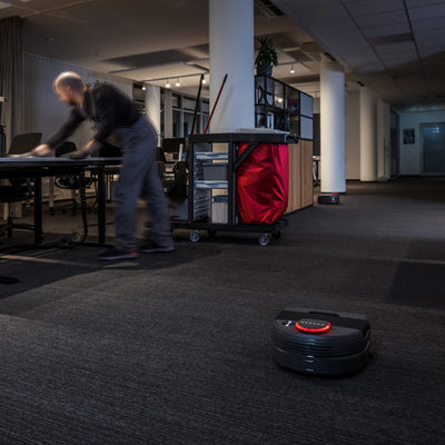 A person tidies an office as a robot vacuum cleans the carpeted floor.