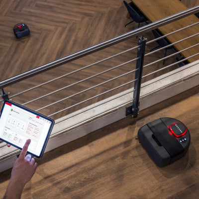 Person uses a tablet to control two robot vacuums on a wooden floor in a modern indoor space.