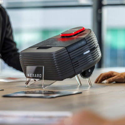 A black Nexaro robot vacuum displayed on a stand, with people sitting around a table nearby.