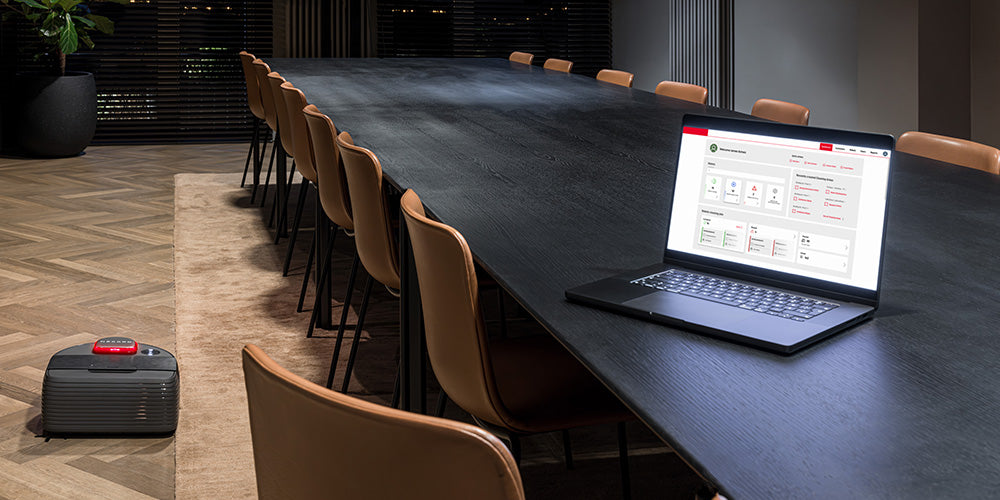 A laptop on a long conference table with a suitcase on the floor nearby in a modern meeting room.
