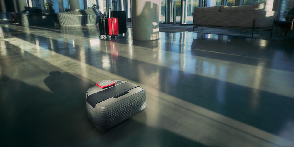 A small robotic suitcase moves across a sunlit lobby floor with two upright suitcases in the background.