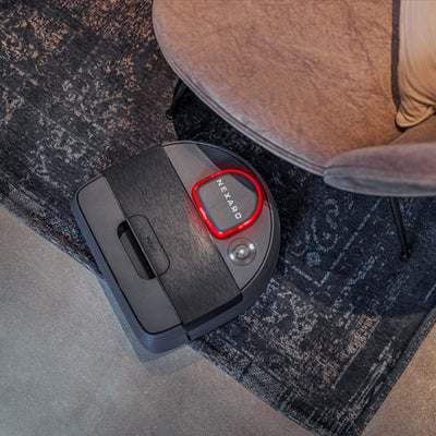 A robot vacuum cleans near a chair on a patterned rug, displaying a red light and NEKARO on top.