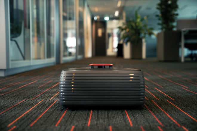 A small, modern robot moves along a carpeted office hallway with glass walls and potted plants.