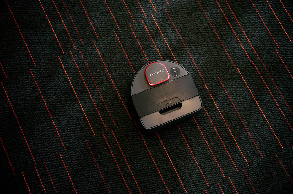 A black Neato robot vacuum cleans a dark carpet with red lines.