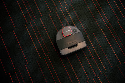 A black Neato robot vacuum cleans a dark carpet with red lines.