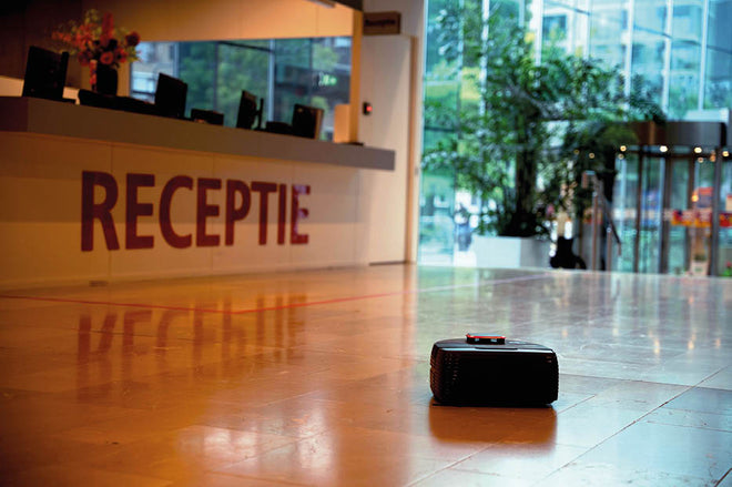 A robot vacuum cleans the shiny lobby floor near a reception desk with RECEPTIE written on it.