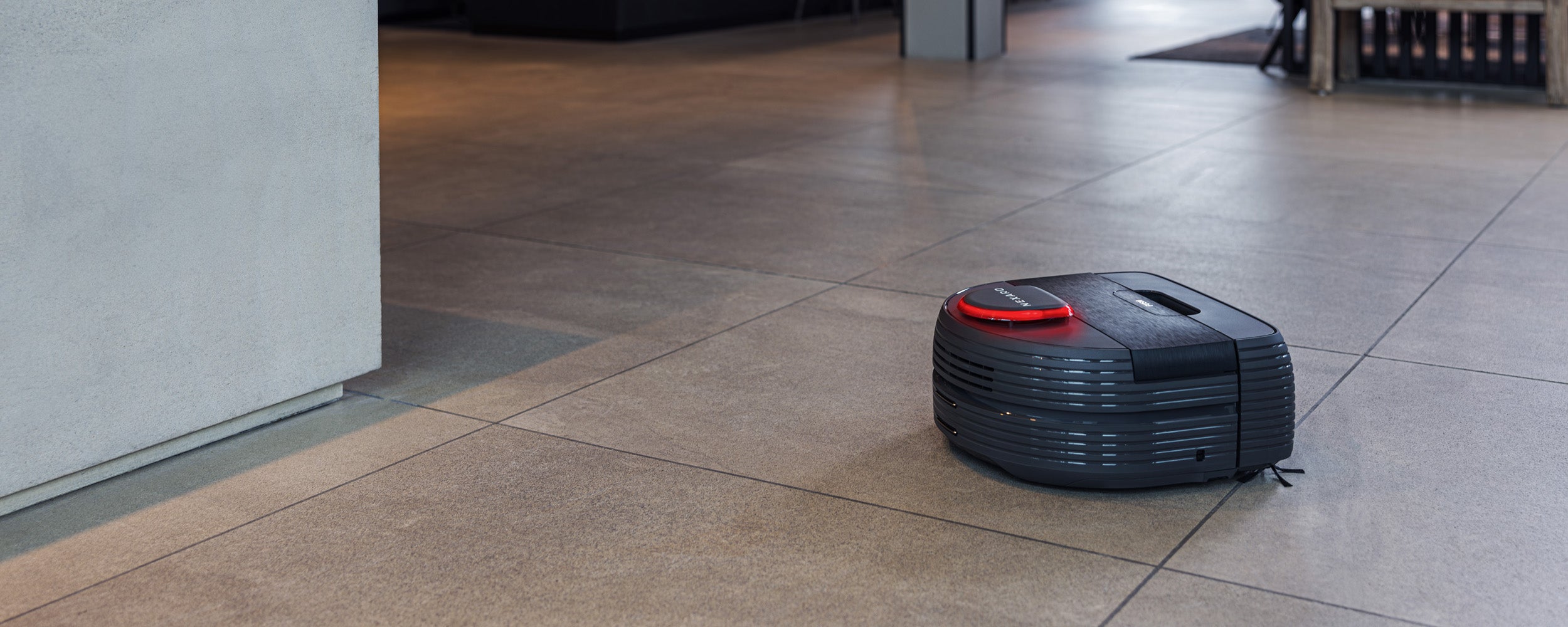 A black robotic vacuum cleaner moves across a tiled floor in a modern home.