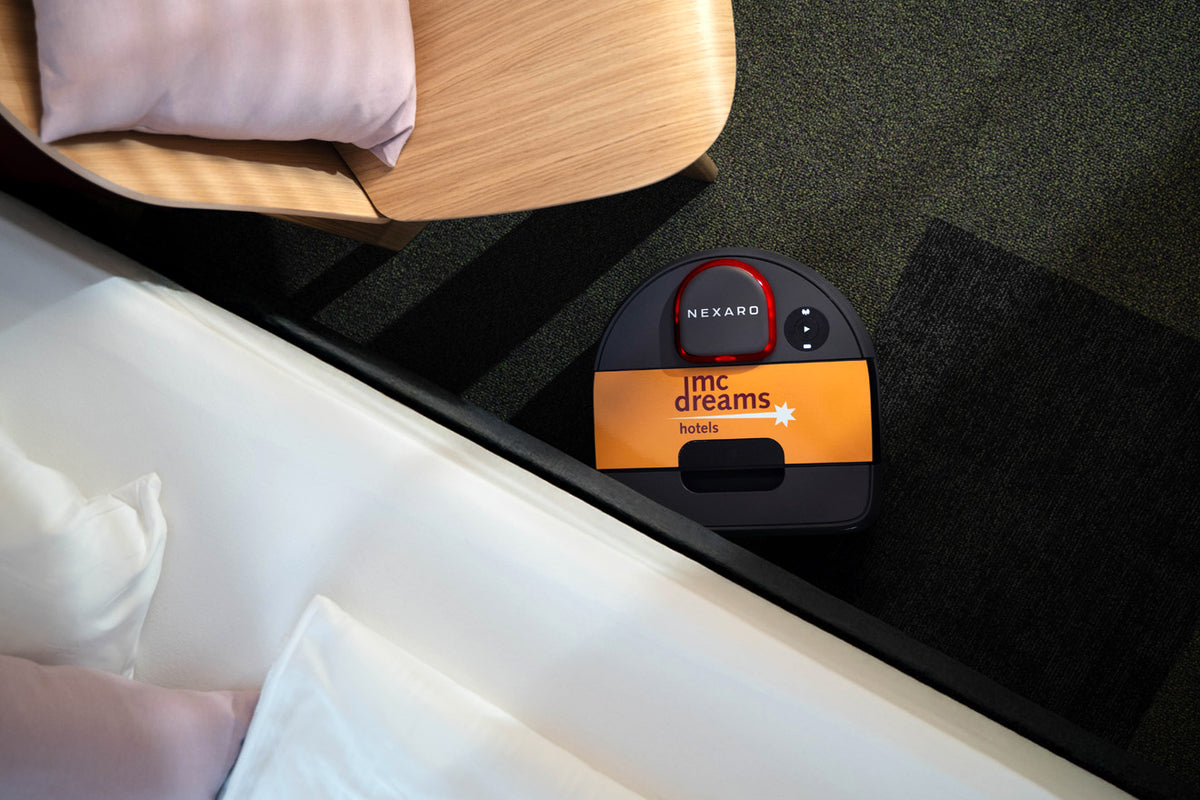 A small delivery robot labeled NEXARD and mc dreams hotels beside a bed and chair.