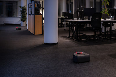 Two robot vacuums clean the floor in a modern, dimly lit office with desks, chairs, and plants.