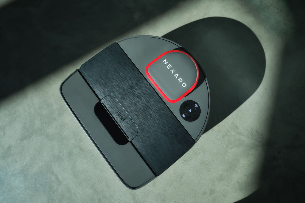 A black and gray Nexaro robotic vacuum sits on a concrete floor, illuminated by sunlight—a striking testament to automatisierte Prozesse and state-of-the-art Automatisierung in modern cleaning solutions.