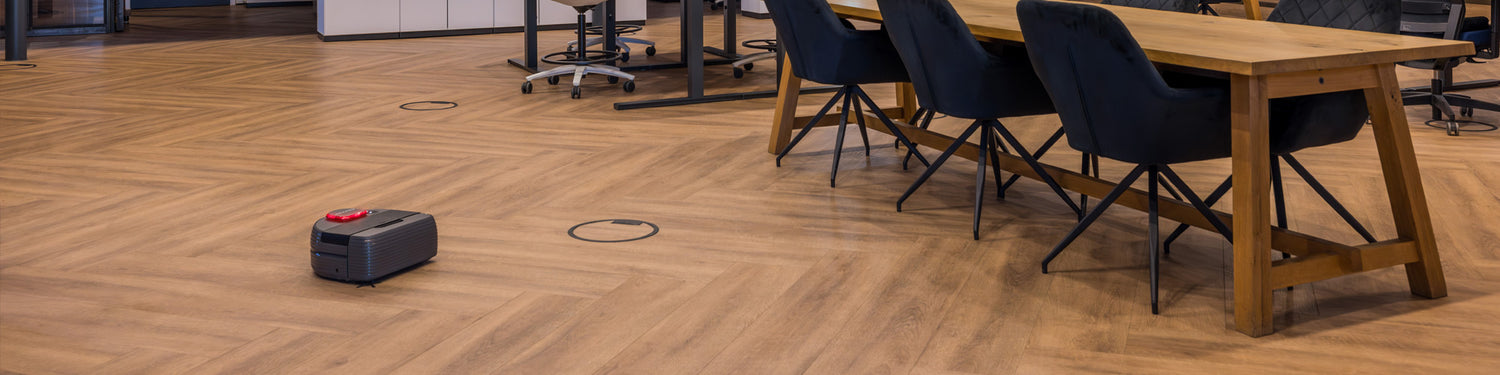 A small robot vacuum cleans the wooden floor near a long table and chairs in a modern office space.