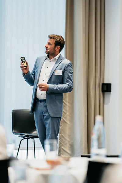Man in a blue suit presenting while holding a device, standing near a chair in a conference room.