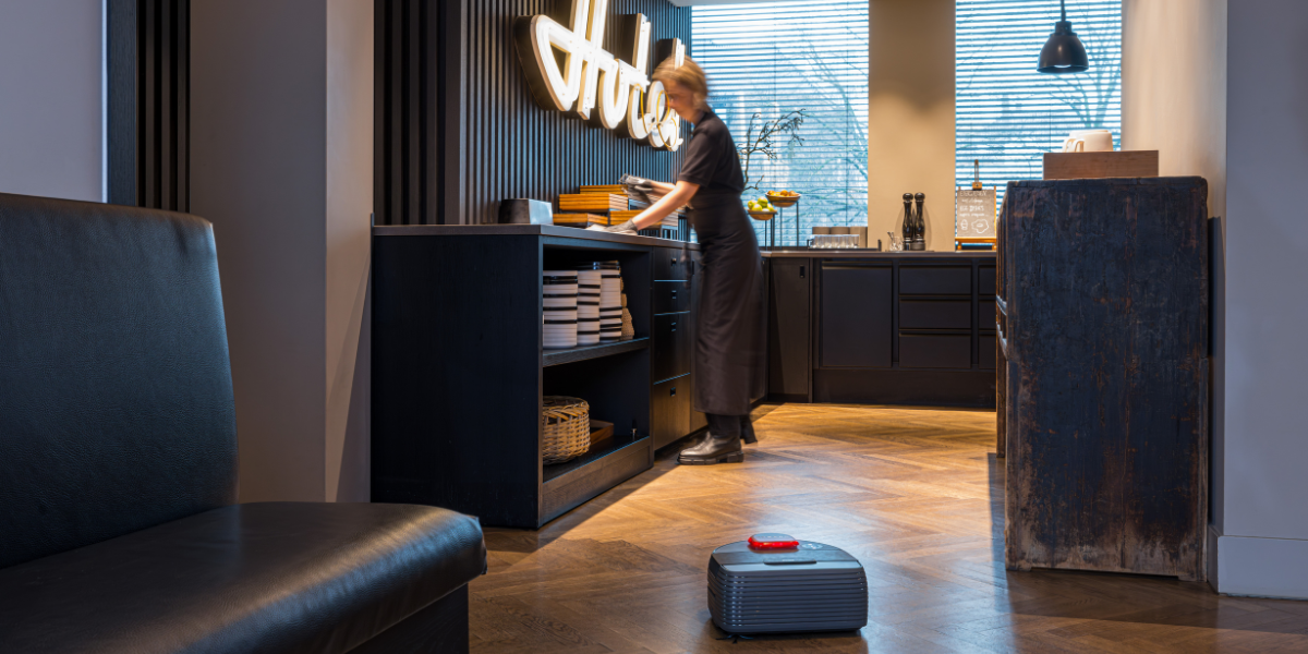 Eine Mitarbeiterin wischt über die Ablagefläche eines Hotel-Büfetts. Neben ihr auf dem Holzboden ist ein Nexaro Saugroboter zu sehen.