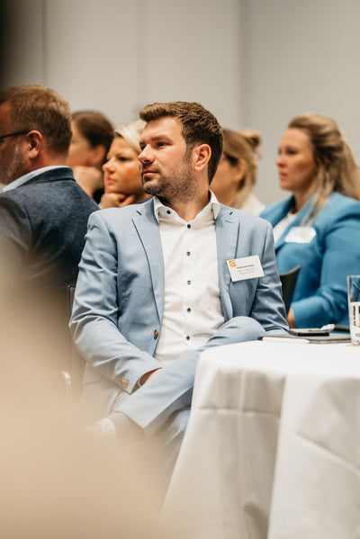 A man in a light blue suit sits at a round table during a conference, surrounded by other attendees.