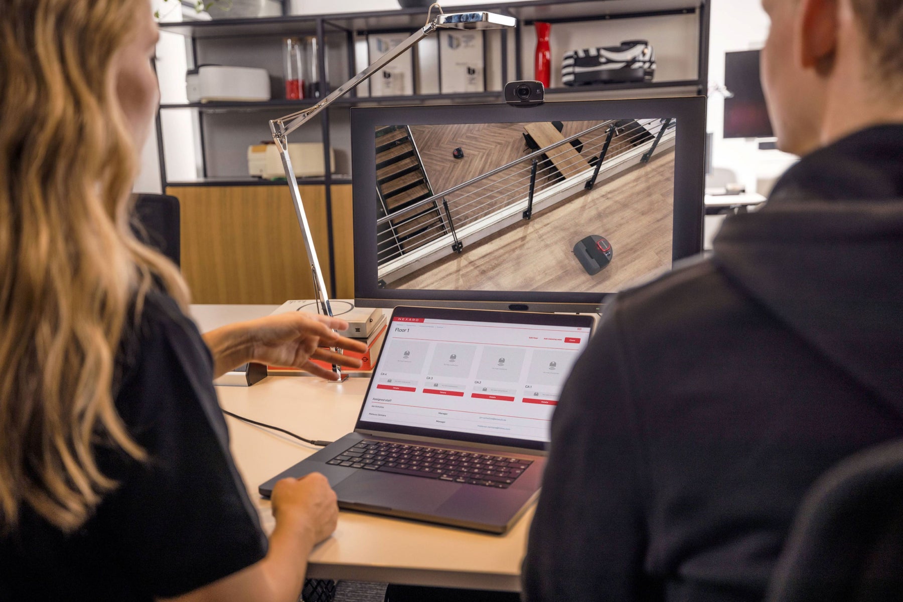 Two people work at a desk with a laptop and monitor displaying a staircase and mouse on a wooden floor.