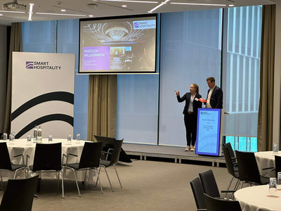 Two speakers present at a Smart Hospitality event in a modern conference room with round tables.