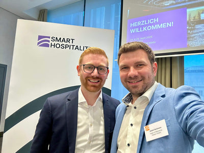 Two men in suits smiling at a Smart Hospitality event, with a welcome sign and presentation screen behind them.